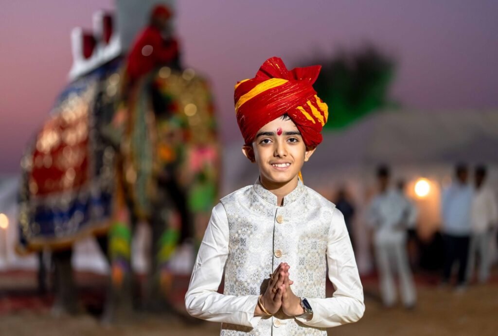 A boy in vibrant Indian clothing poses outdoors at an evening event with an elephant.